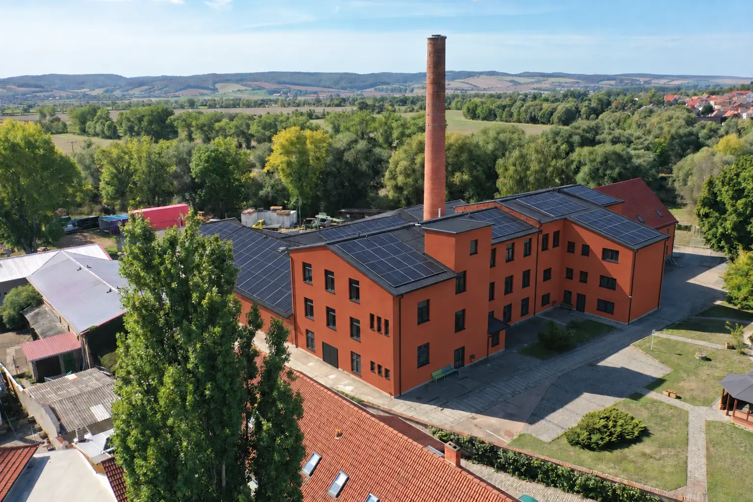 KI-Gestaltungsidee für Hauptbau und Halle: rotbrauner Putz, schwarze Fenster und Trapezblechdächer mit Photovoltaik